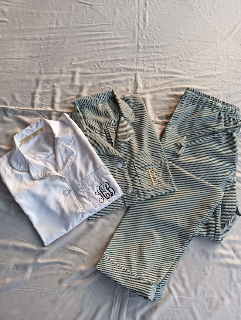May include: Three sets of pajamas are displayed on a gray surface. One set is white, and the other two are green and white striped. The white set has a monogram on the cuff. The green set has a monogram on the pocket. The pajama pants have a drawstring.
