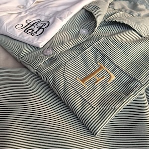 May include: Two button-down shirts, one white and one green striped, are neatly folded. The white shirt features black embroidered initials, while the green shirt has a gold embroidered initial. The shirts are made of a soft, likely cotton, fabric.