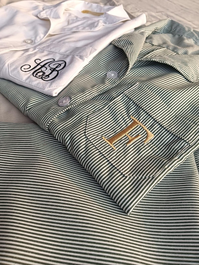 May include: Two button-down shirts, one white and one green striped, are neatly folded. The white shirt features black embroidered initials, while the green shirt has a gold embroidered initial. The shirts are made of a soft, likely cotton, fabric.