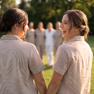 May include: Two people wearing brown and white striped short-sleeved shirts, holding hands, and smiling. Other people are in the background, wearing similar outfits. The setting is outdoors, with green grass and trees.