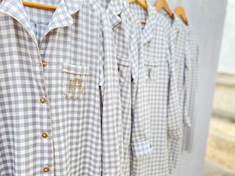 May include: Five gray and white gingham shirts with embroidered initials on the pocket. The shirts are hanging on wooden hangers.