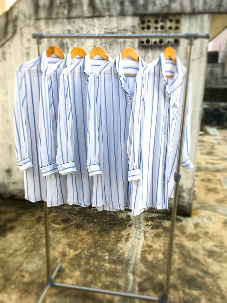 May include: Five long-sleeved button-down shirts hanging on a metal rack. Each shirt is white with vertical blue stripes, featuring a notched collar, a front button placket, and cuffs on the sleeves. The shirts are displayed side by side.
