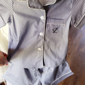 May include: Blue and white striped pyjama set with white trim. The set includes a short-sleeved button-down shirt with a chest pocket and shorts. The shirt pocket has a black embroidered initial. The tag reads "What a lovely".