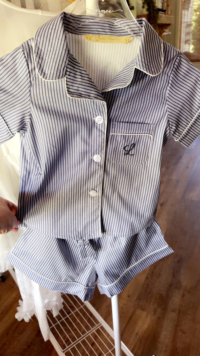May include: Blue and white striped pyjama set with white trim. The set includes a short-sleeved button-down shirt with a chest pocket and shorts. The shirt pocket has a black embroidered initial. The tag reads "What a lovely".