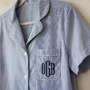 May include: Blue and white striped pajama top with short sleeves, a notched collar, and a chest pocket. The pocket features a monogram in navy blue thread. The shirt is buttoned and hangs on a wooden hanger.