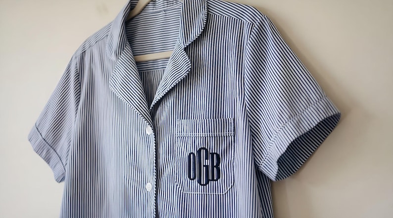 May include: Blue and white striped pajama top with short sleeves, a notched collar, and a chest pocket. The pocket features a monogram in navy blue thread. The shirt is buttoned and hangs on a wooden hanger.