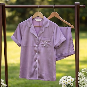 Pode incluir: Um conjunto de pijama lavanda composto por uma camisa de manga curta com botões, gola de lapela e bolso no peito, acompanhado por calções a condizer. A camisa apresenta debrum branco e uma letra H bordada a branco no bolso.