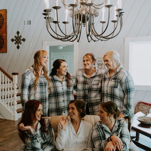 Peut inclure: Un groupe de sept femmes dans une pièce avec un lustre. Six des femmes portent des pyjamas en plaid assortis, et une femme porte une robe blanche avec les initiales "CH" brodées sur le devant. Elles sourient toutes et se regardent.