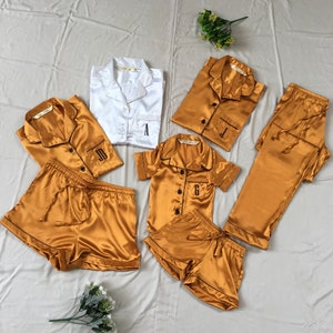 May include: A set of matching pajamas in gold satin with black trim. The pajamas include a short-sleeved shirt, shorts, and pants. Each piece is personalized with a different letter: A, J, and G.
