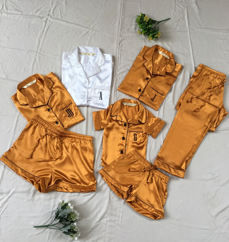 May include: A set of matching pajamas in gold satin with black trim. The pajamas include a short-sleeved shirt, shorts, and pants. Each piece is personalized with a different letter: A, J, and G.