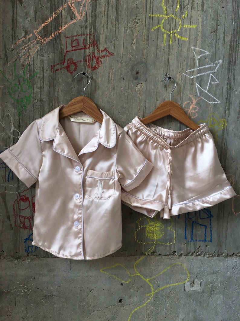 Puede incluir: Un conjunto de pijama de dos piezas en color melocot&oacute;n claro. La parte superior tiene mangas cortas, un frente abotonado y un ribete blanco. Los pantalones cortos tienen una cintura el&aacute;stica y un ribete blanco. El conjunto est&aacute; hecho de sat&eacute;n.