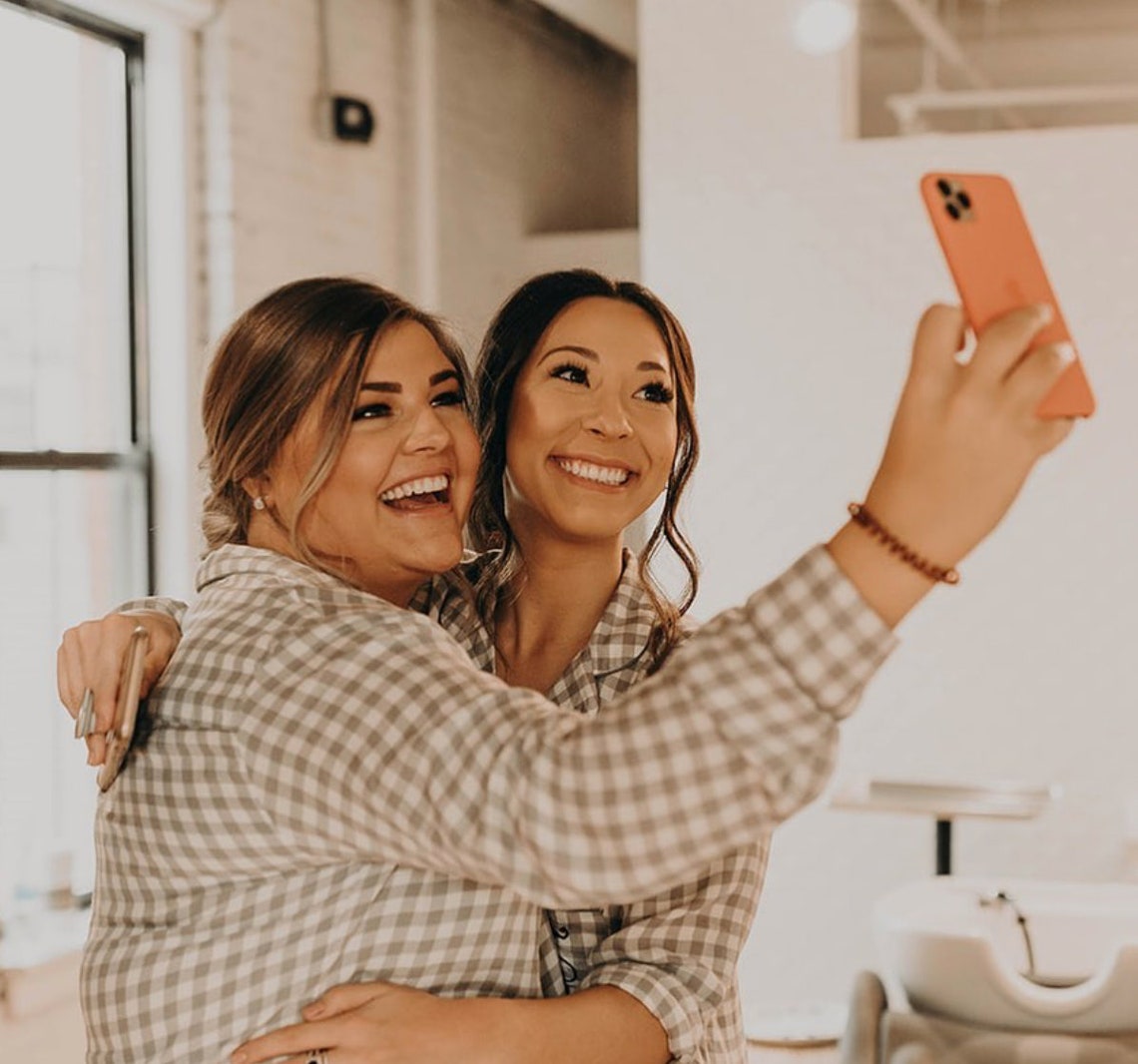 May include: Two people wearing plaid shirts are taking a selfie with a pink phone. One person is laughing and the other is smiling.