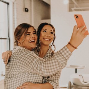 May include: Two people in matching plaid shirts are taking a selfie. The person in the back is holding a pink phone and smiling. The person in the front is laughing and has their arm around the other person.