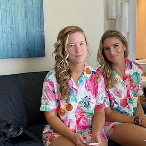 May include: Two people wearing matching floral print pyjamas with short sleeves and a button-down front. The pyjamas have a pocket on the left side with the initials "SML" embroidered on them.