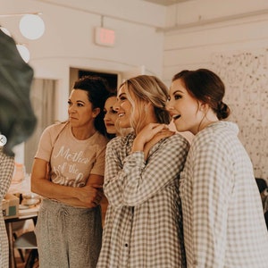 May include: A group of women in a room, smiling and looking at something off-camera. One woman is wearing a pink shirt with the text "Mother of the Bride". The other women are wearing gray and white plaid shirts.