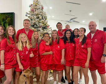 May include: A group of people wearing matching red pajama sets pose for a photo in front of a decorated Christmas tree. The pajamas have white trim and personalized initials. A dog is also present.