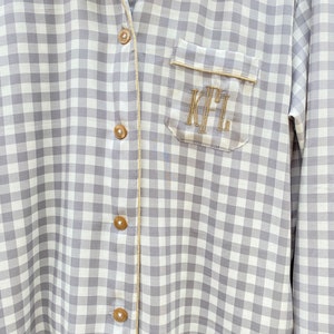 May include: A gray and white gingham button-down shirt with a gold trim and a pocket on the left side. The pocket has the initials "K.F.L." embroidered in gold thread.