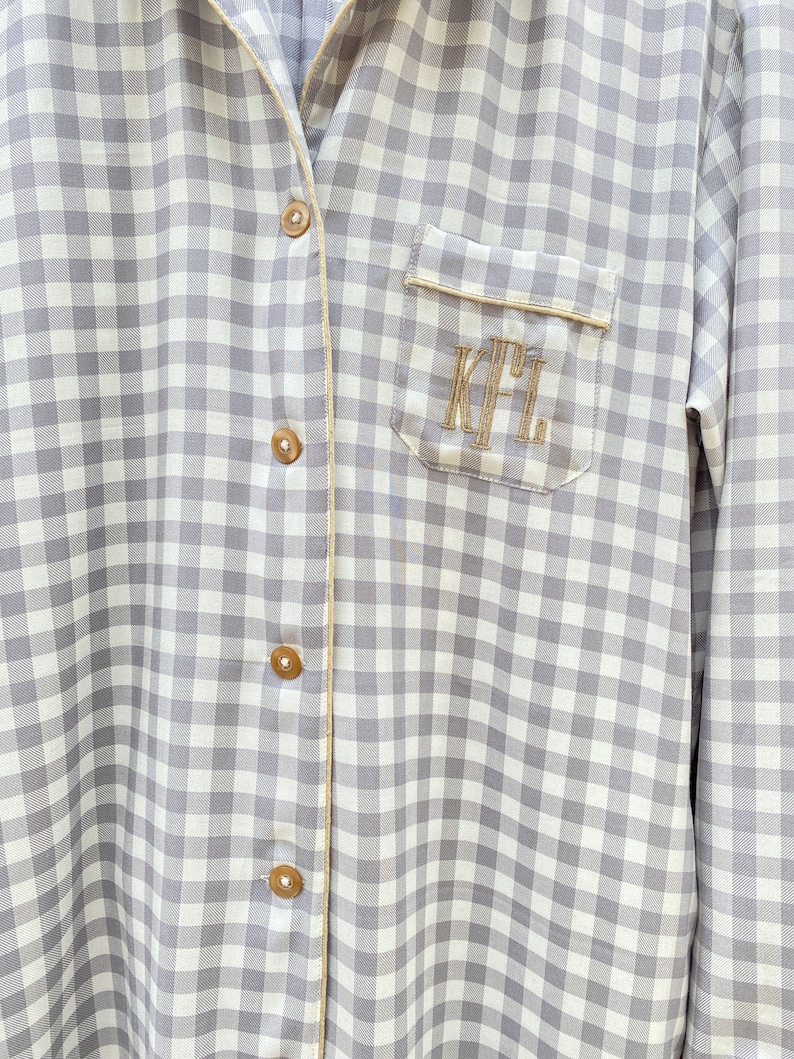 May include: A gray and white gingham button-down shirt with a gold trim and a pocket on the left side. The pocket has the initials "K.F.L." embroidered in gold thread.