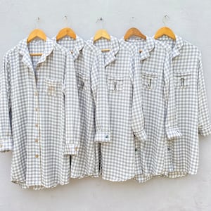 May include: Five gray and white gingham shirts with black trim and embroidered initials. The shirts are hanging on wooden hangers against a white wall.