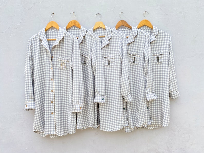 May include: Five gray and white gingham shirts with black trim and embroidered initials. The shirts are hanging on wooden hangers against a white wall.