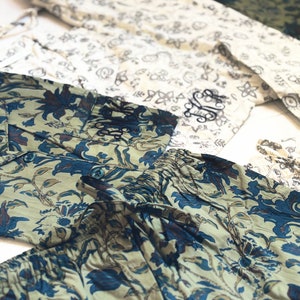May include: Several pairs of patterned pajama pants in various colors. One pair features a blue and beige floral design, another has a white and black floral pattern, and a third is olive green. The pants are laid out on a white surface.