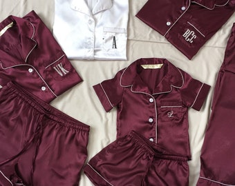 May include: Burgundy and white satin pajama sets with white piping. The sets include button-down shirts and shorts, some with embroidered initials. One white shirt is also visible. The pajamas are arranged on a white sheet.