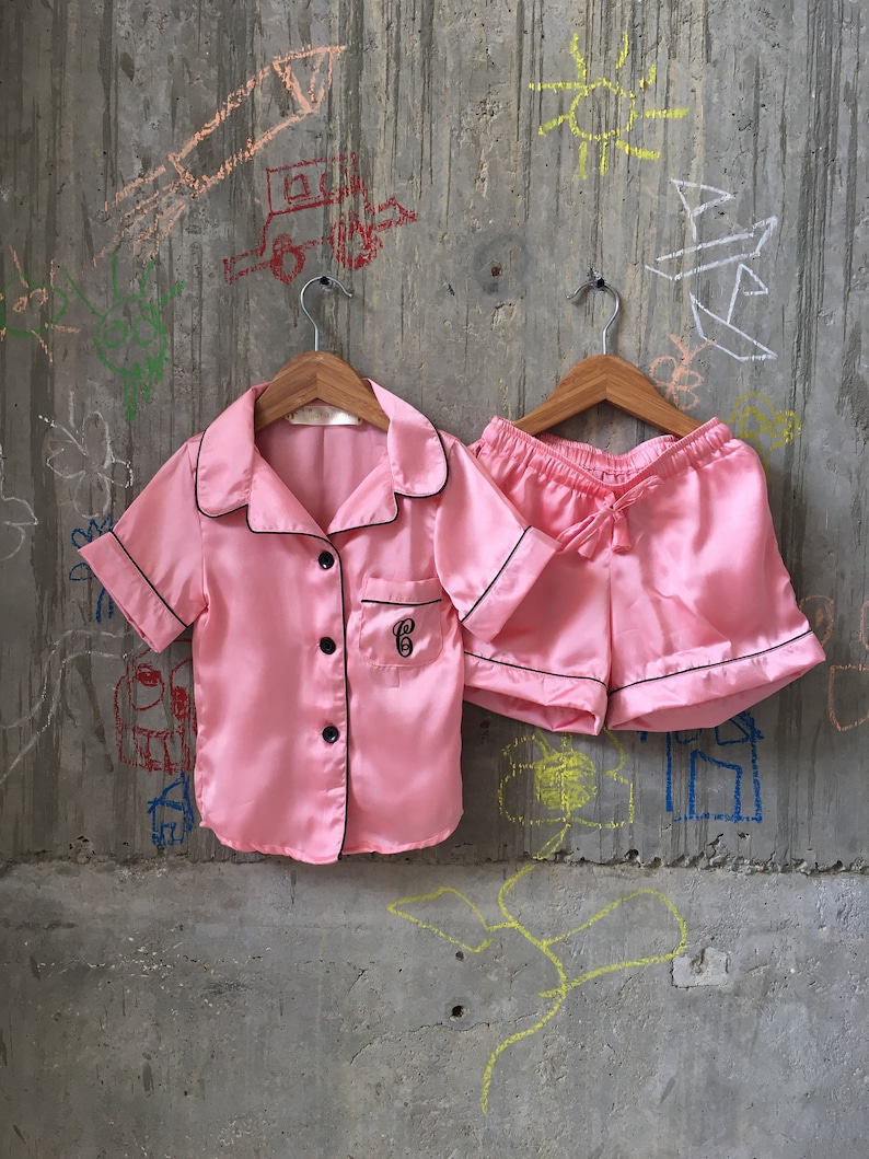 Puede incluir: Un conjunto de pijama de sat&eacute;n rosa con ribetes negros. La parte superior tiene un frente abotonado y una letra "G" bordada en negro en el pecho. Los pantalones cortos tienen una cintura con cord&oacute;n.