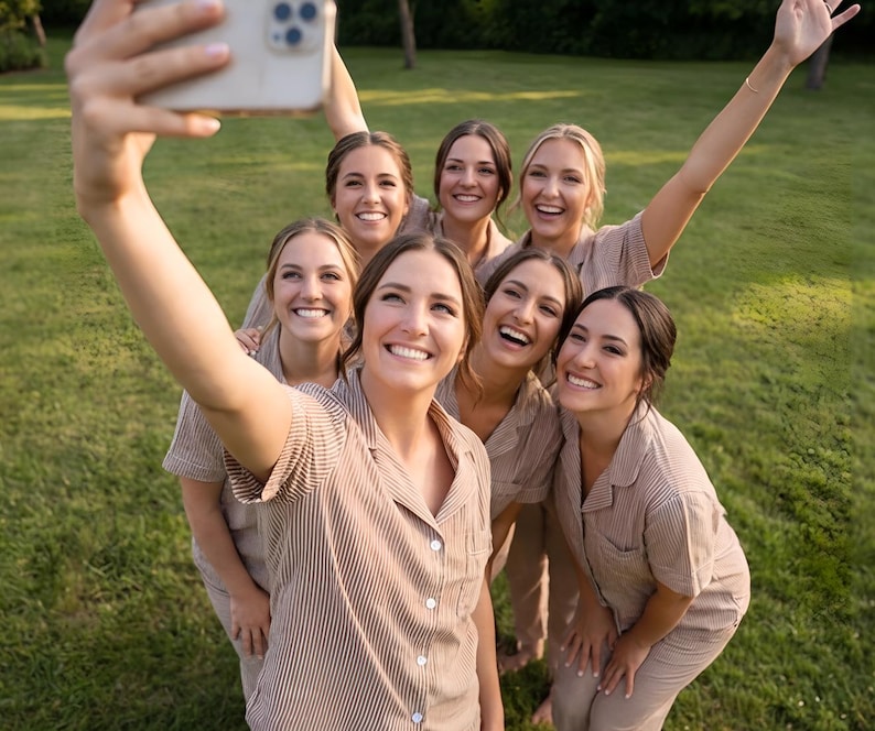 May include: A group of seven women wearing matching beige striped pajama sets, smiling and taking a selfie outdoors on a sunny day. The women are standing on a green grassy lawn.