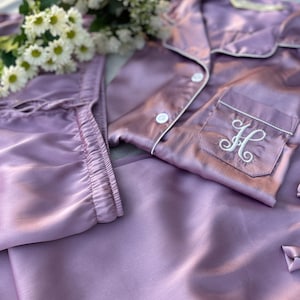 May include: Lilac-colored satin pajama set with white piping and a pocket embroidered with the letter "H". The set includes a button-down shirt and pants. A bouquet of white flowers rests near the pajamas.