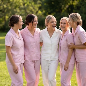 May include: Five people wearing matching pink and white pajama sets, standing on a grassy lawn. The pink sets have a striped pattern. One person wears a white pajama set. All are smiling and laughing.