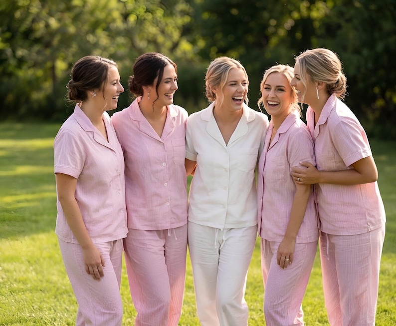 May include: Five people wearing matching pink and white pajama sets, standing on a grassy lawn. The pink sets have a striped pattern. One person wears a white pajama set. All are smiling and laughing.