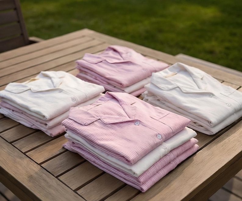 May include: Four stacks of folded pajama sets on a wooden table. The sets include white and pink striped or solid-colored button-down shirts and matching bottoms. The pajamas are neatly arranged, suggesting a focus on comfort and relaxation.