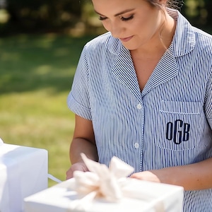 May include: A blue and white striped pajama shirt with short sleeves and a breast pocket featuring the embroidered initials "OGB". The shirt is buttoned and worn by a person. White gift boxes with bows are in the foreground.