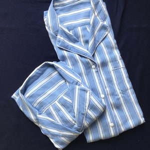 May include: A light blue and white striped pajama set featuring a long-sleeved button-down shirt with a notched collar and a chest pocket, paired with matching pajama bottoms. The fabric has a textured finish and white circular buttons.