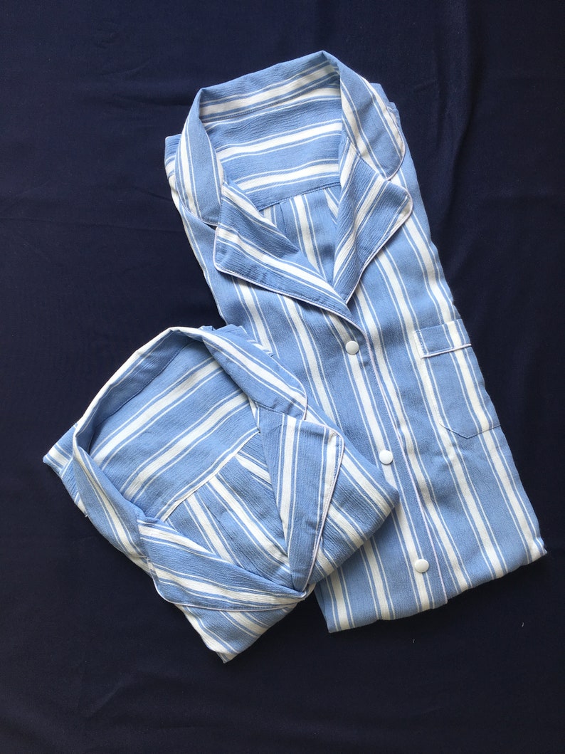 May include: A light blue and white striped pajama set featuring a long-sleeved button-down shirt with a notched collar and a chest pocket, paired with matching pajama bottoms. The fabric has a textured finish and white circular buttons.