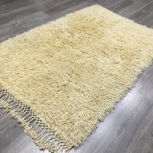 May include: A cream-colored shaggy rug with a fringe border. The rug is made of soft, fluffy fibers and has a luxurious feel.