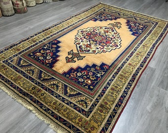 May include: A traditional Persian rug with a beige background and intricate floral patterns in shades of blue, red, and gold. The rug has a fringed edge and is laid out on a wooden floor.