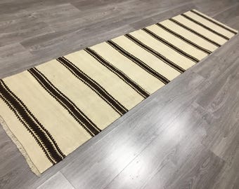 May include: A long, narrow, cream-colored rug with brown stripes. The rug has a fringe on both ends.