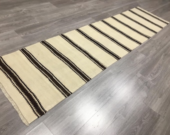 May include: Cream-colored runner rug with dark brown stripes. The rug has a rectangular shape and a fringed edge. The stripes are evenly spaced across the length of the rug. The rug is made of a woven material.