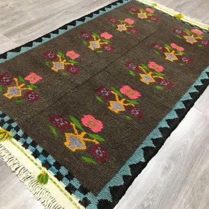 Peut inclure: Un tapis brun avec un motif géométrique de fleurs colorées. Le tapis a une bordure de franges blanches avec des accents bleus, noirs et jaunes.