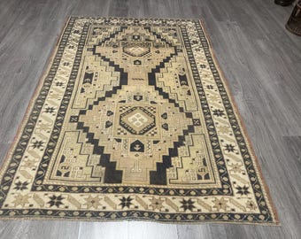 May include: A rectangular rug with a beige and black geometric pattern. The rug has a central design with black and beige shapes, framed by a border of repeating star and geometric patterns. It is placed on a grey wooden floor.