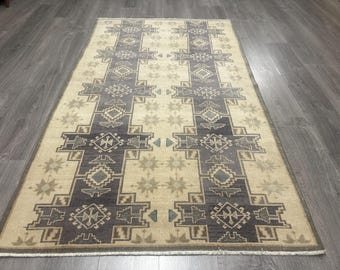 May include: A rectangular rug in beige with a geometric design in grey and tan. The pattern includes repeating cross-like shapes and star motifs, creating a symmetrical design. The rug has a border and is placed on a wooden floor.