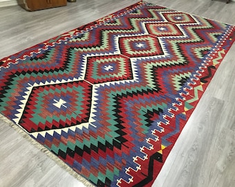 May include: A traditional Turkish rug with a geometric pattern in shades of red, blue, green, and black. The rug has a fringed edge and is woven with intricate details.