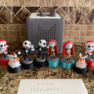 May include: A collection of nine Nightmare Before Christmas figurines, including Jack Skellington, Sally, Oogie Boogie, and Zero. The figurines are arranged in a row on a countertop, with a gray speaker in the background. The figurines are all wearing their signature costumes and have a festive Christmas theme.