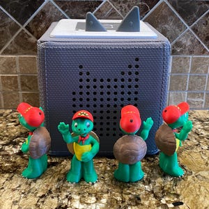 Könnte beinhalten: Ein dunkelgrauer Lautsprecher mit Haifischflossen-Akzenten oben und vier grünen Schildkrötenfiguren mit roten Hüten und braunen Panzer. Die Schildkröten sind in verschiedenen Posen, einige winken.