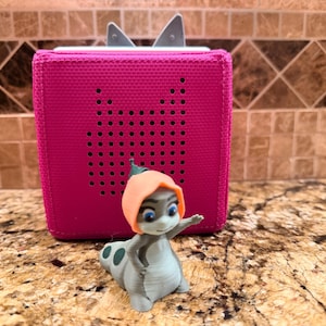 May include: A bright pink square speaker with a perforated cat-ear design on the front, positioned behind a small, light gray figurine of a creature with a peach-colored hood, blue eyes, and a spotted tail, standing in a waving pose.