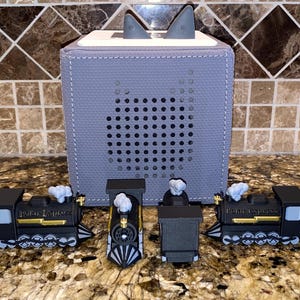 May include: A gray fabric speaker with a perforated front and two gray ears on top. Four toy train cars are in front of the speaker. The train cars are black with gold accents and the words "Polar Express".