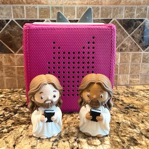 May include: A magenta fabric speaker with a gray shark fin antenna is behind two small figurines of Jesus holding a book. The figurines have brown hair and beards, and are wearing white robes. The speaker has a perforated design.