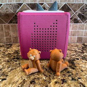 May include: Two brown teddy bears sit in front of a pink speaker with a white top and grey ear-like accents. The speaker has a grid of small holes on the front.