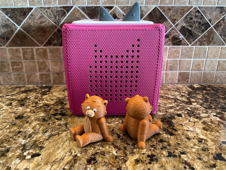 May include: Two brown teddy bears sit in front of a pink speaker with a white top and grey ear-like accents. The speaker has a grid of small holes on the front.